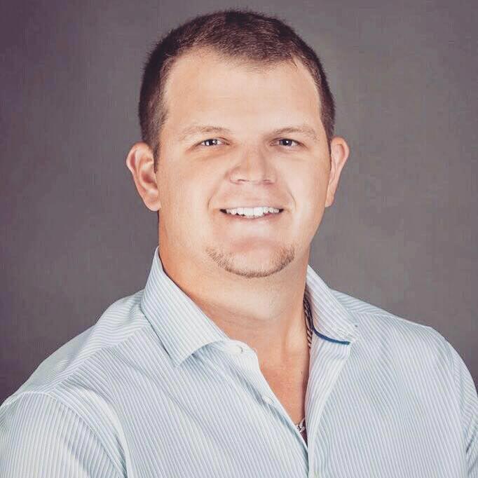 The image shows a man wearing a light blue shirt with a darker blue tie, smiling at the camera.